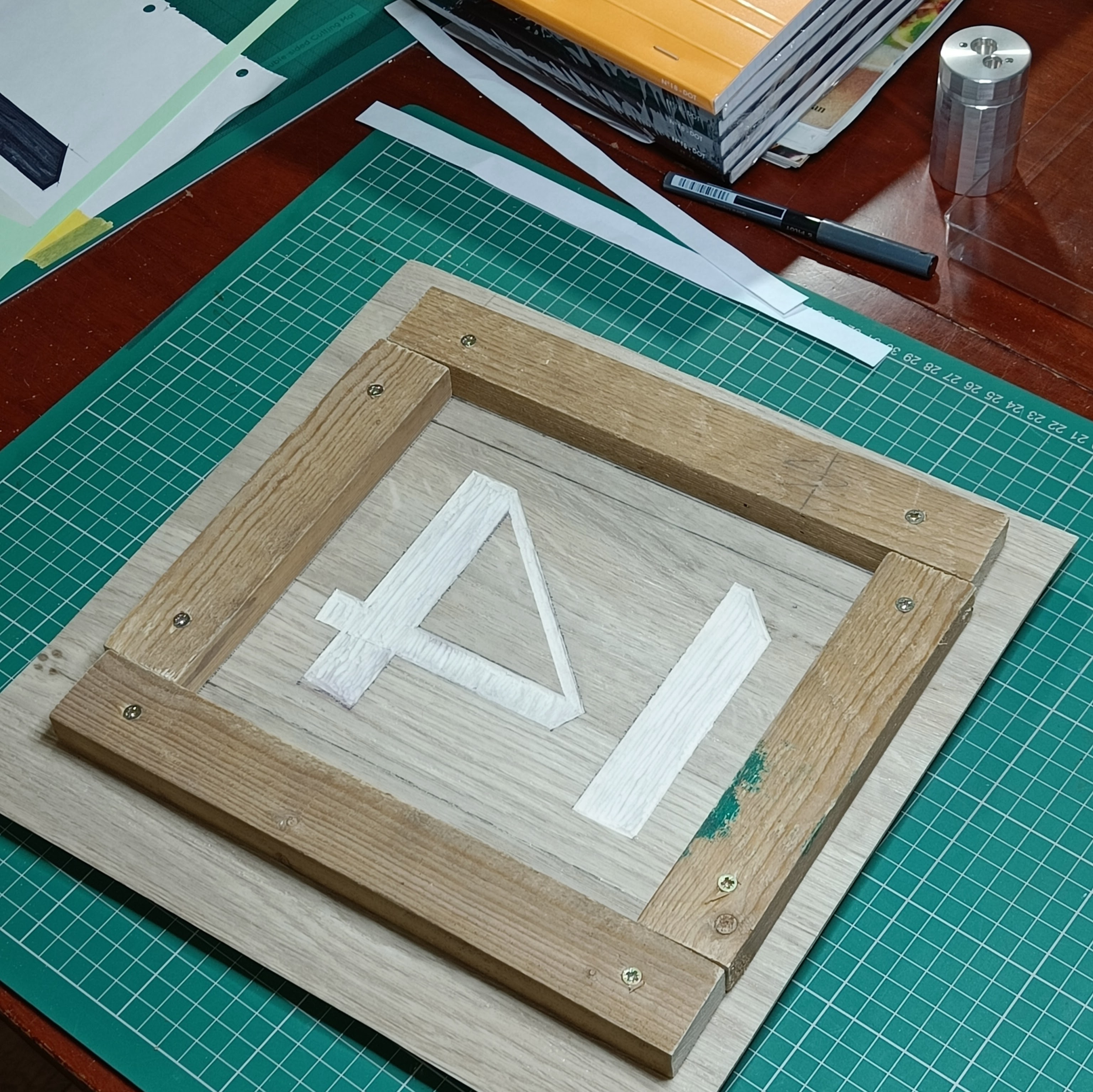 a crude wooden frame is screwed onto a piece of cut
  bathroom vinyl (showing the number 14). The vinyl is slightly
  lifted off from the cutting mat, suggesting the piece of OSB
  behind. A few items lay about on the table: a Pilot V5 pen, an
  expensive pencil sharpener made from aircraft aluminium.